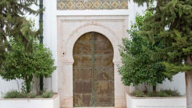 Large studded wooden double door in a pale stone arch, topped by a patterned tile frieze and flanked by trees.