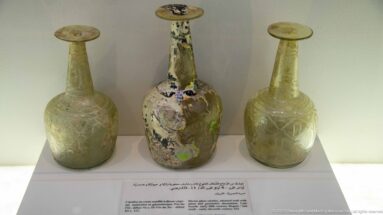 Three ancient long-necked glass bottles with round bodies and flared rims, including one heavily weathered iridescent vessel at center.