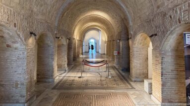 Long brick barrel-vaulted gallery with repeated side arches, protected mosaic floors, spotlights, and a bright doorway at the far end.