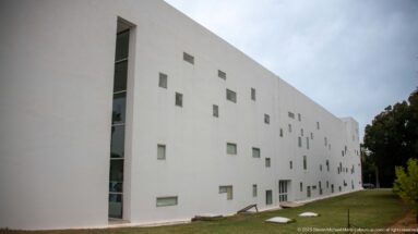 Long white modern museum wing with scattered square and rectangular windows, a vertical glazed corner, and a narrow lawn alongside.