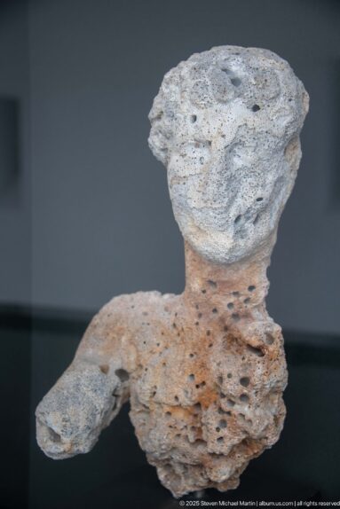 Small human-shaped artifact heavily covered by pale mineral encrustation, with head, torso, and one arm visible.