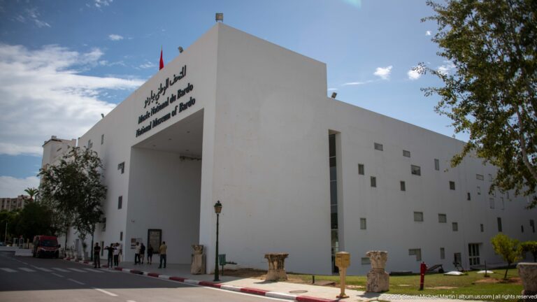 Modern white Bardo Museum entrance with multilingual signage, small square windows, and stone antiquities lining the approach.