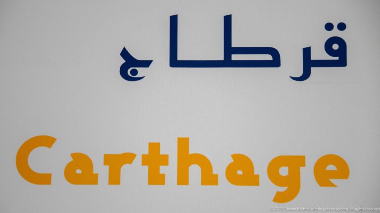 White gallery wall with the word Carthage in ochre beneath large dark blue Arabic lettering.