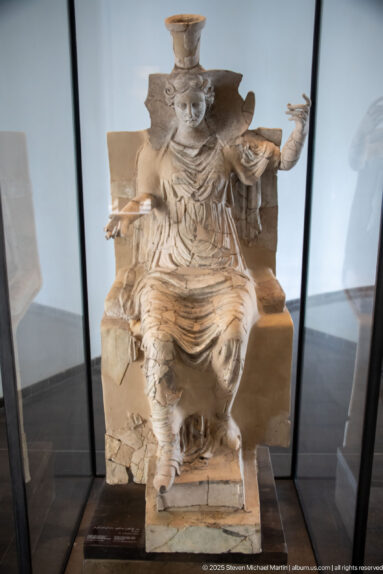 Terracotta enthroned woman wearing a tall headdress and layered garments, with one arm extended.