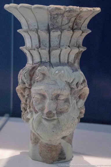 Anthropomorphic terracotta vessel shaped as a bearded head with traces of red pigment and a wide flaring neck.