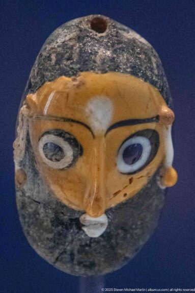 Small dark glass head-shaped vessel with large blue-and-white eyes, orange brow, ears, and a top opening.