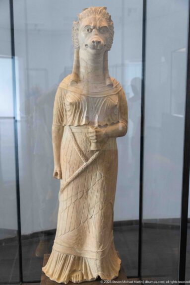 Standing stone female figure with stylized hair, mantle, and long dress incised with a diamond pattern.