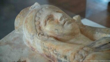 Close view of a reclining carved stone head with curled hair resting against a funerary support.