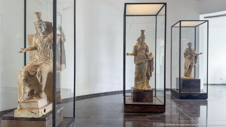 Bright white museum gallery with several large ancient figures displayed individually in tall glass vitrines.