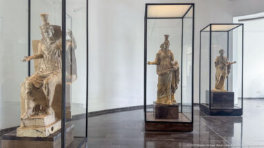 Bright white museum gallery with several large ancient figures displayed individually in tall glass vitrines.