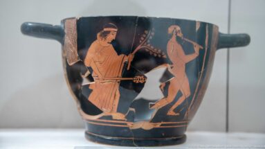 Black-ground ceramic cup with two reddish standing male figures and a projecting side handle.