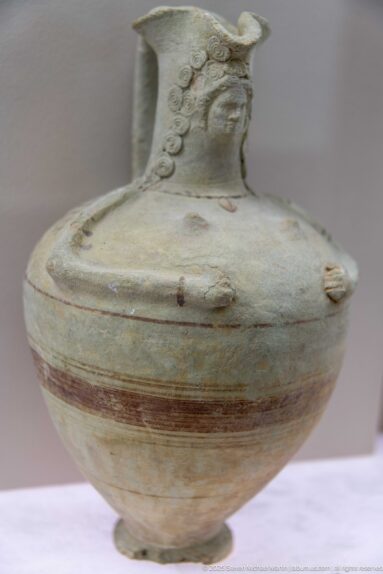 Pale squat ceramic flask with a narrow neck, small foot, applied round ornaments, and faded horizontal bands.