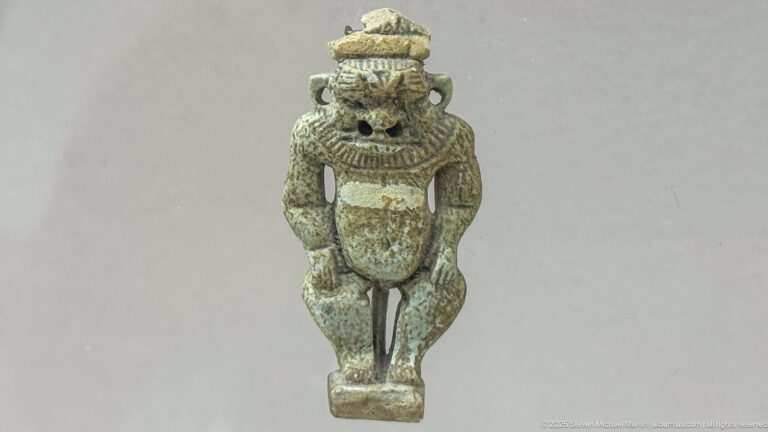 Small greenish Bes-type figurine with a leonine face, high headdress, rounded body, and frontal stance.