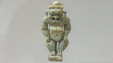 Small greenish Bes-type figurine with a leonine face, high headdress, rounded body, and frontal stance.