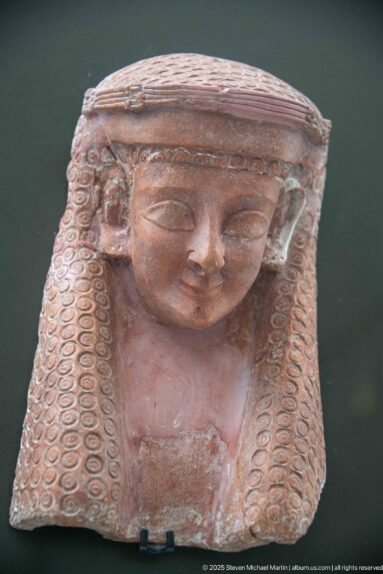 Terracotta female head or bust with a flat headdress, elongated eyes, and rows of tightly curled side locks.