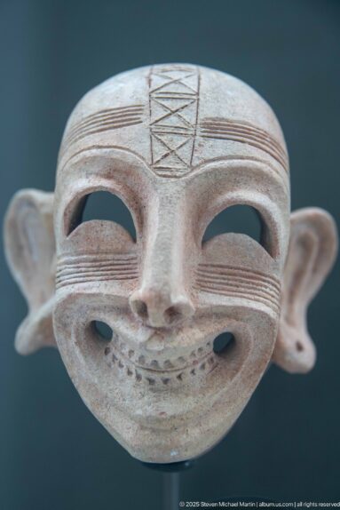 Terracotta mask with hollow eyes, broad grin, projecting ears, cheek lines, and a hatched geometric forehead motif.
