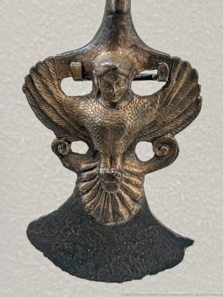 Small bronze winged figure with a humanlike head, broad feathered wings, openwork sides, and fan-shaped tail.