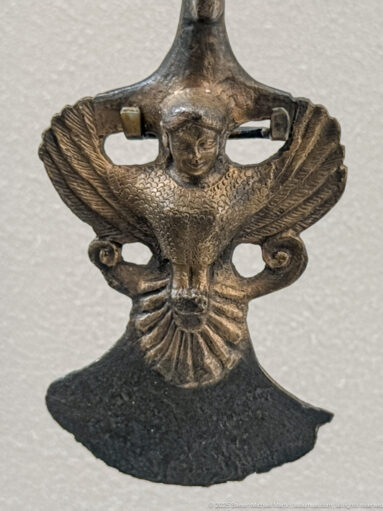 Small bronze winged figure with a humanlike head, broad feathered wings, openwork sides, and fan-shaped tail.
