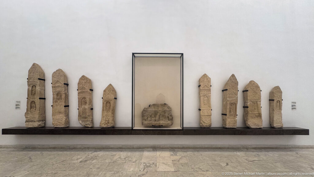 Row of narrow carved stone stelae displayed along a museum wall on both sides of a central case.