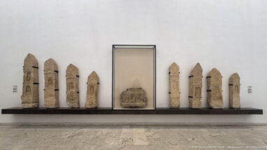 Row of narrow carved stone stelae displayed along a museum wall on both sides of a central case.