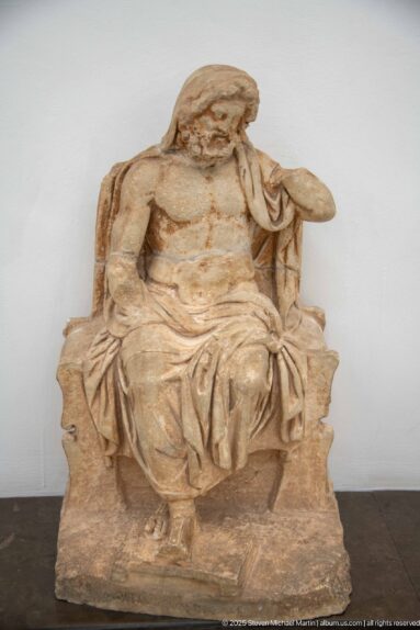 Carved stone seated male figure with draped clothing, exposed chest, head covering, and deeply folded garment across the lap.