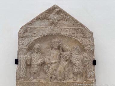 Gabled stone relief with a central standing female figure, smaller attendants, animals, and dense carved ornament.