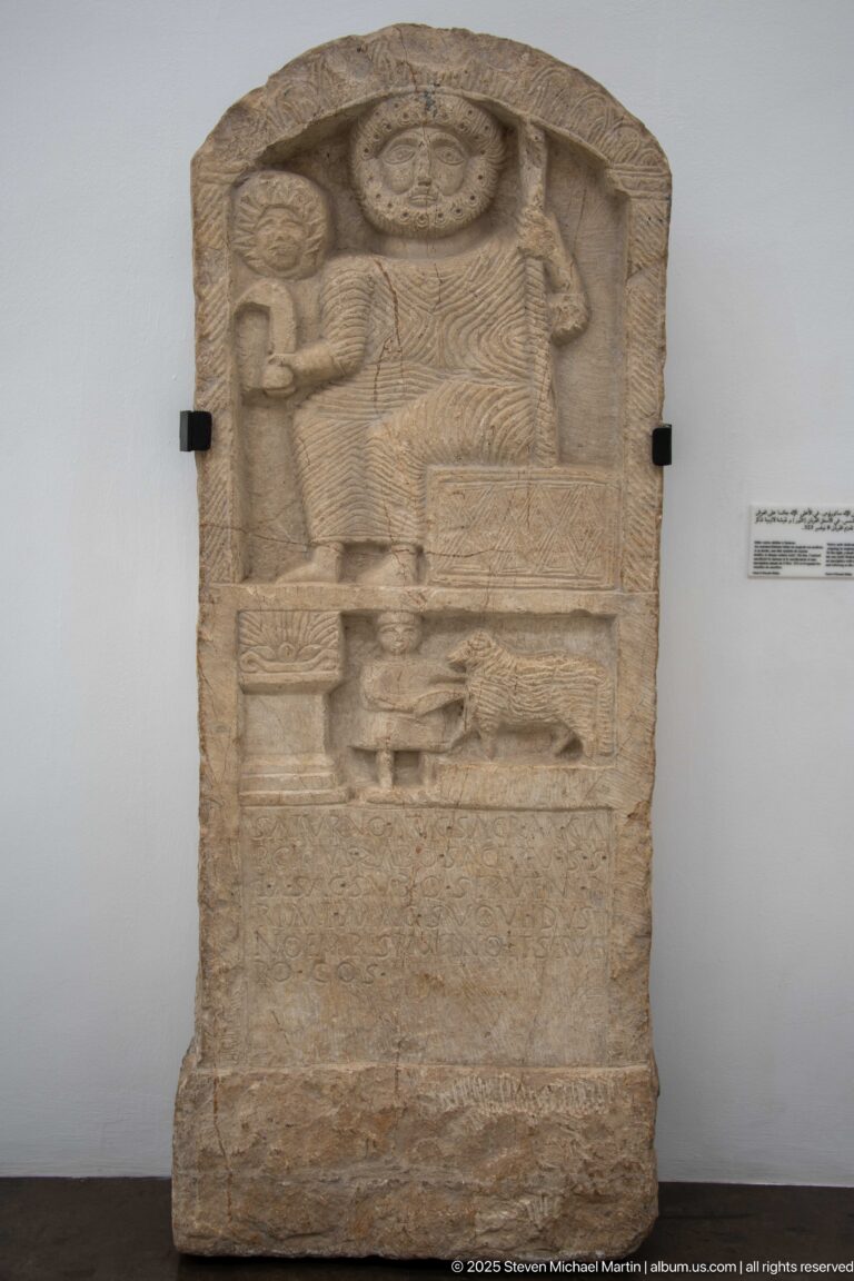 Arched stone stele with a large central figure beneath a radiating disk and smaller carved figures in surrounding registers.