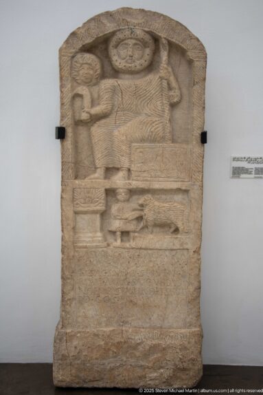 Arched stone stele with a large central figure beneath a radiating disk and smaller carved figures in surrounding registers.