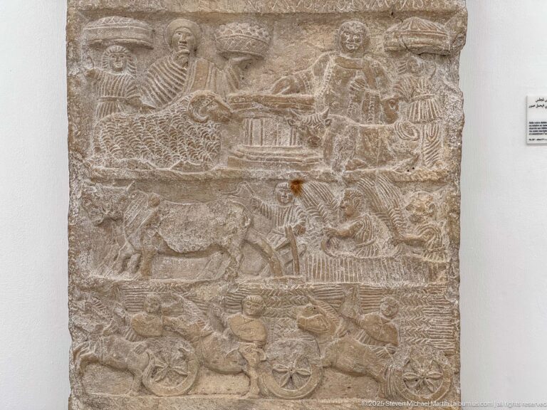 Carved stone relief with several registers containing people, animals, a central table scene, and wheeled carts.