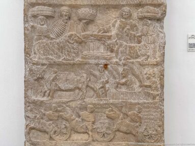 Carved stone relief with several registers containing people, animals, a central table scene, and wheeled carts.