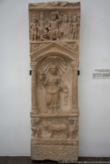 Tall carved stone monument with a robed standing figure in a framed niche and a crowded relief register above.