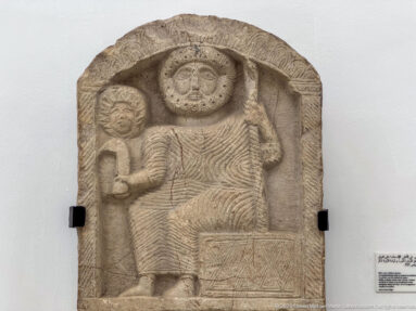 Arched stone stele with a large seated figure, radiating disk above, raised arm, and decorated rectangular support.