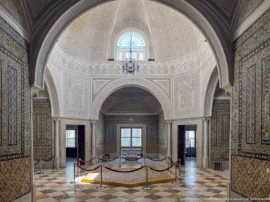 Ornate Bardo Museum interior with carved white plaster, tiled walls, broad arches, marble columns, and patterned floor.