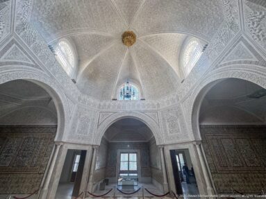 Upward view of white carved-plaster vaults and arches with high windows and a gilded ceiling boss.