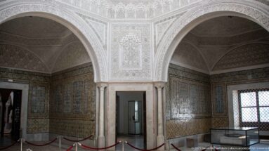 Corner of the Bardo Museum interior with tiled walls, carved-plaster arches, marble columns, and a latticed window.
