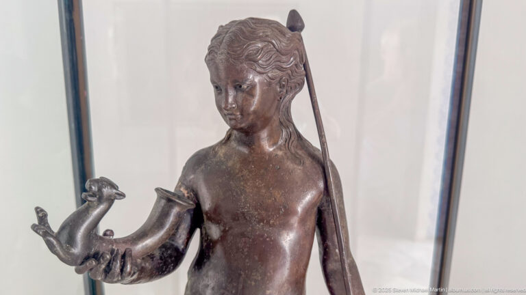 Bronze female statuette with wavy hair, a long support behind her shoulder, and a small animal resting on one hand.