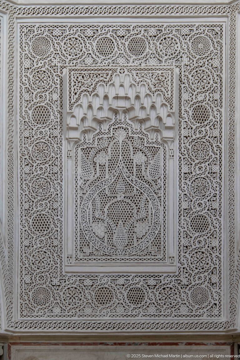 White carved-plaster panel with dense geometric and vegetal patterns surrounding a recessed muqarnas-like central niche.