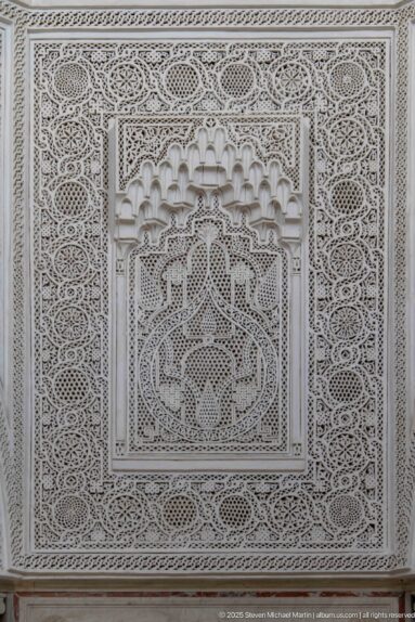 White carved-plaster panel with dense geometric and vegetal patterns surrounding a recessed muqarnas-like central niche.