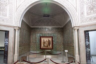 Framed figural mosaic displayed at the end of a tiled palace alcove beneath a carved-plaster arch.