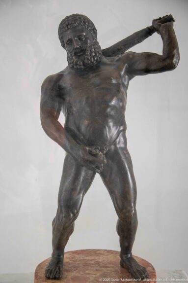 Bearded nude bronze male figure standing wide-legged and holding a long club or staff diagonally behind his shoulders.