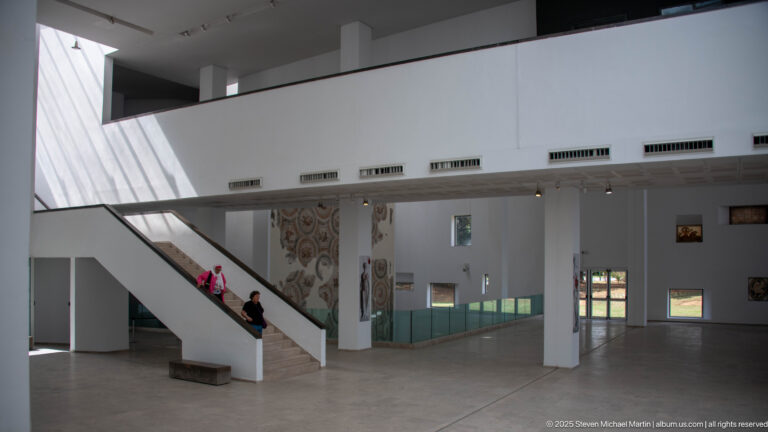 White museum gallery with diagonal staircase, glass display cases, wall-mounted mosaic, and two visitors.