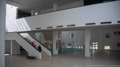White museum gallery with diagonal staircase, glass display cases, wall-mounted mosaic, and two visitors.