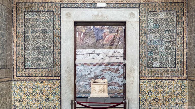 Framed archaeological display set into a richly patterned tiled wall, with several mosaic fragments above a small stone object.