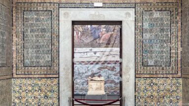 Framed archaeological display set into a richly patterned tiled wall, with several mosaic fragments above a small stone object.