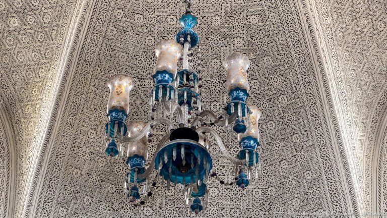 Blue-and-clear glass chandelier suspended beneath a densely carved white plaster ceiling.
