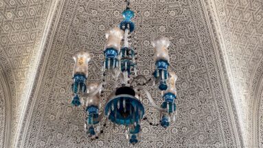 Blue-and-clear glass chandelier suspended beneath a densely carved white plaster ceiling.