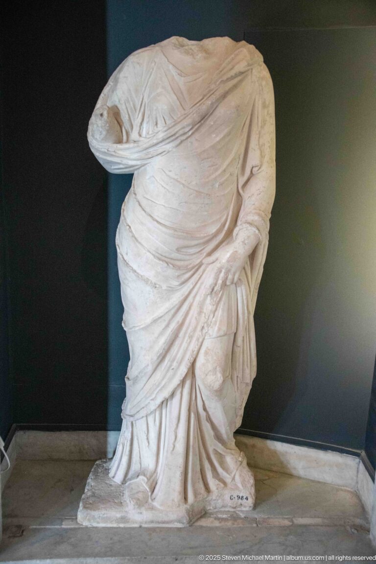 Headless marble standing figure wrapped in deeply folded drapery, with one surviving hand gathering the garment at the hip.