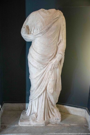 Headless marble standing figure wrapped in deeply folded drapery, with one surviving hand gathering the garment at the hip.