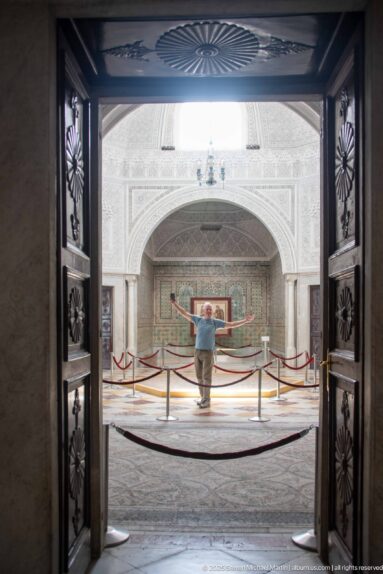 Open carved wooden doors framing a visitor and mosaic display in a bright vaulted gallery beyond.