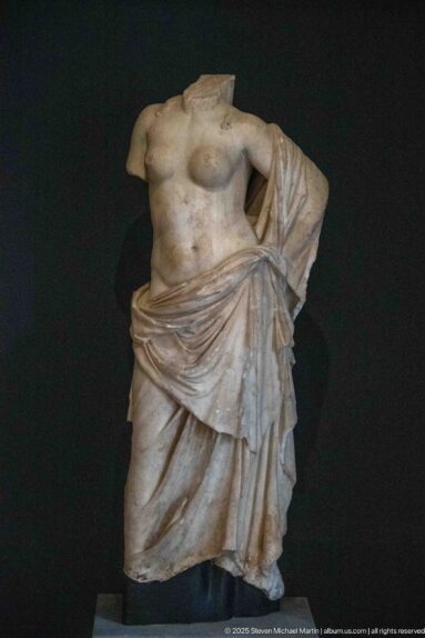 Headless marble female torso with one breast exposed and heavy drapery wrapped diagonally around the hips.